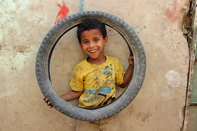 A smiling boy in a yellow T-shirt looks out through the centre of a tyre held against a wall.
