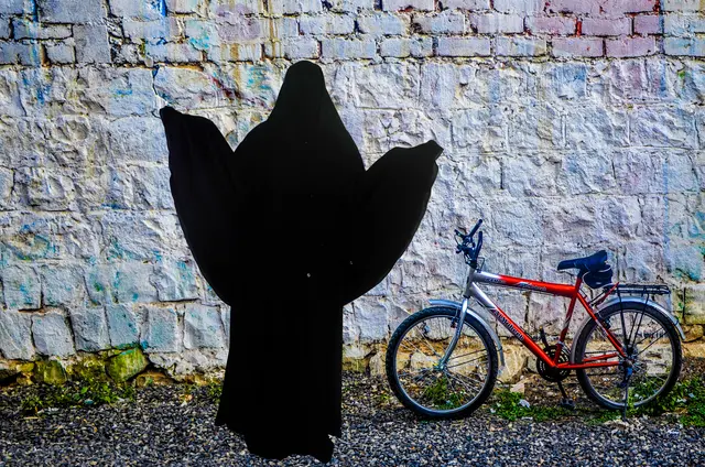 A figure in a black abaya stands with arms spread against a whitewashed wall, beside a red bicycle.