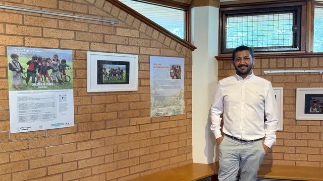 A man in a white shirt stands beside a Children of Yemen poster, a framed photograph and a text panel on a brick wall.