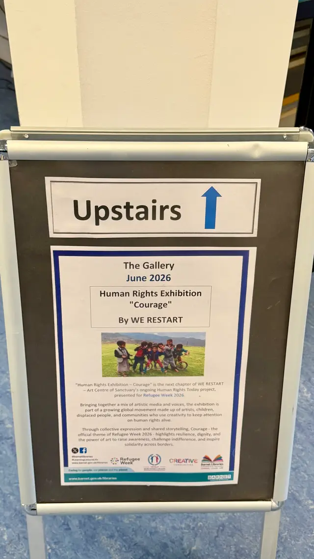 A library sign pointing upstairs to the gallery: ‘Human Rights Exhibition “Courage” by WE RESTART’, June 2026.