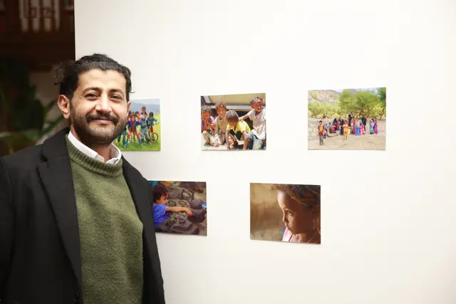 A man smiles beside a wall of small colour photographs of children.