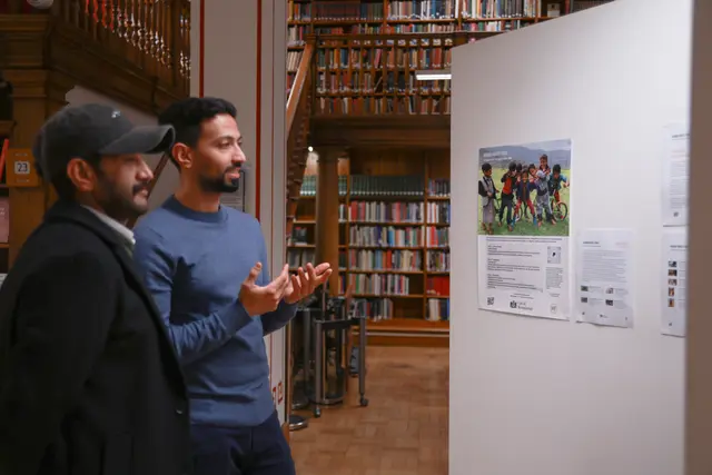 Two men talk beside the exhibition display, with library bookshelves behind them.