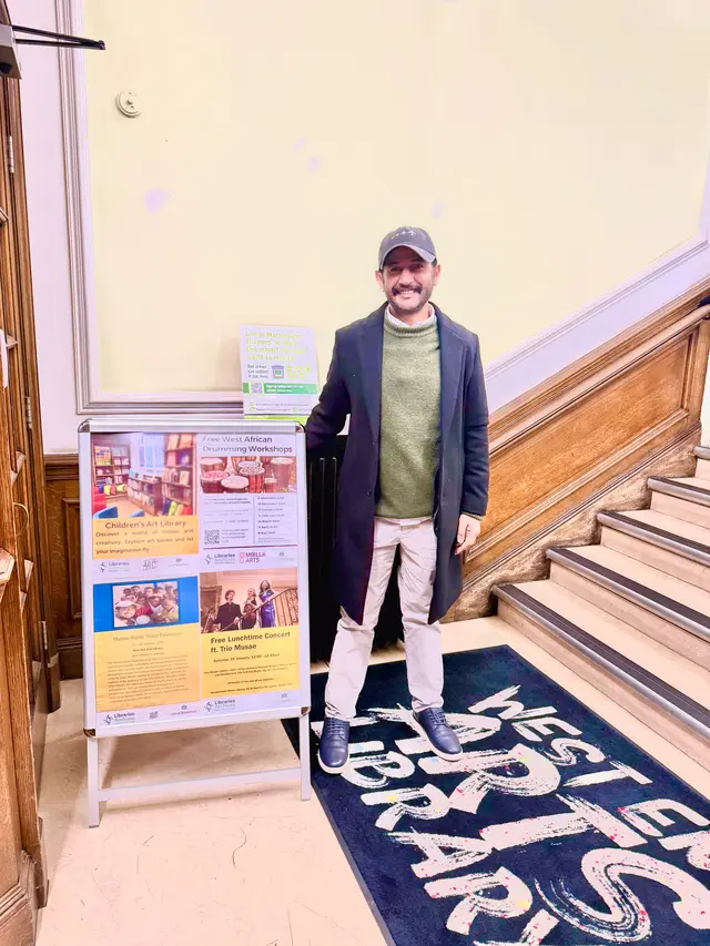 An exhibition poster on an easel at the foot of a library staircase, with a man standing beside it.