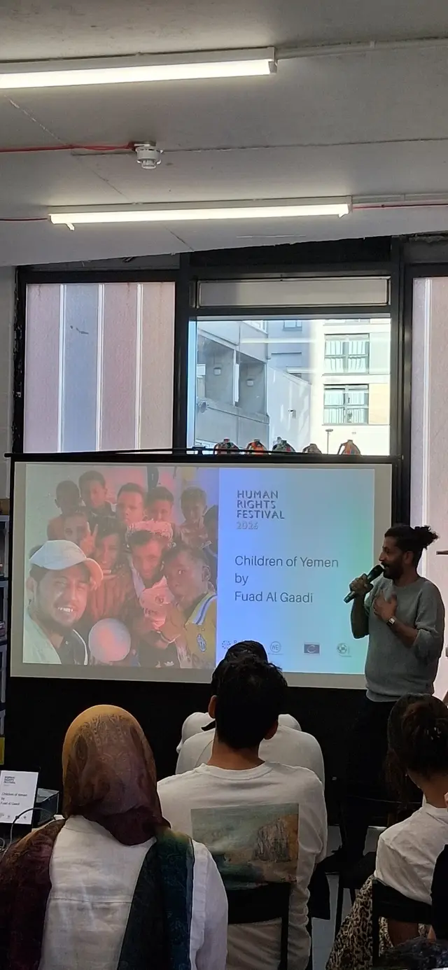 A speaker with a microphone stands beside a screen reading ‘Children of Yemen by Fuad Al Gaadi’, Human Rights Festival 2026, in front of a seated audience.