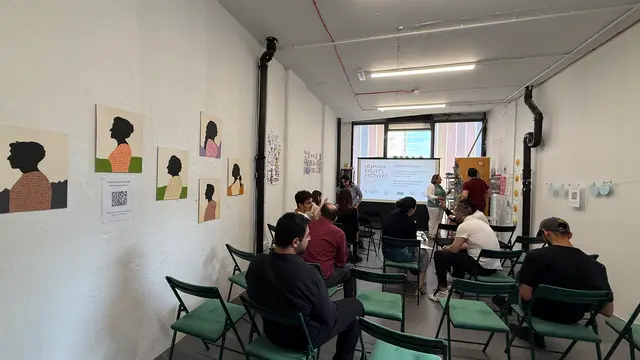 An audience seated on folding chairs in a white-walled room, facing a screen.