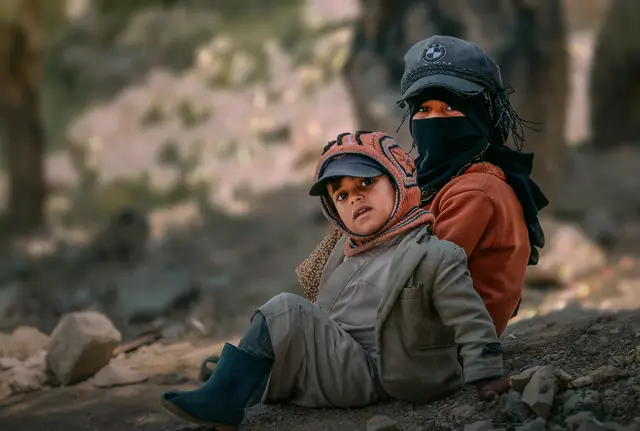 Two children in knitted hats sit close together on rocky ground.