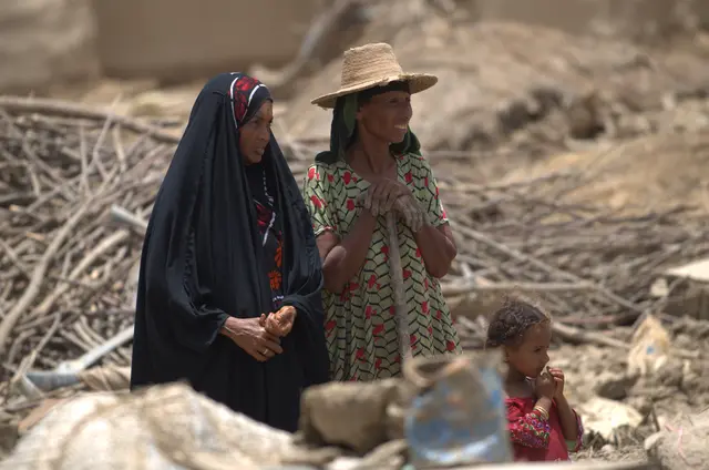 Two women, one in a black abaya and one in a wide straw hat, stand among dry branches while a small child sits nearby.