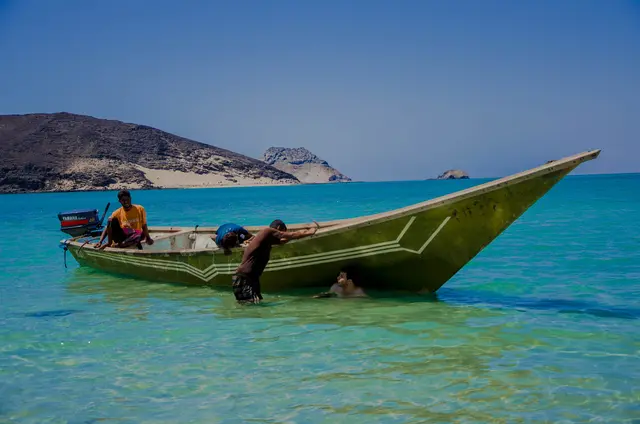 A wooden boat in clear turquoise water near the shore, with two people beside it.